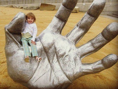 A little girl sitting on a giant hand rising from the ground sculpture at the National Children’s Mu...