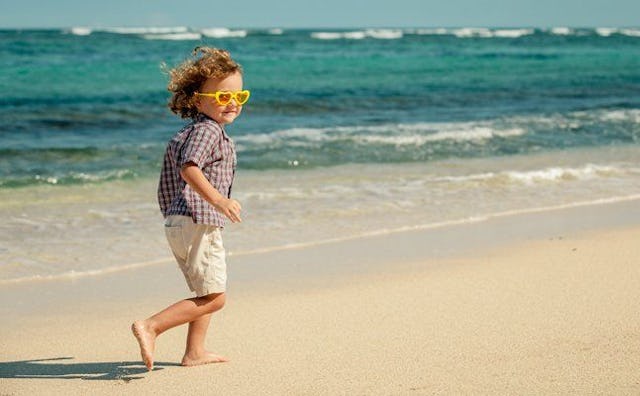 A young boy with long brown curly hair and sunglasses running on the beach near the sea on a sunny d...