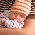 A new dad wearing a brown and white striped t-shirt holding his baby that’s wearing a striped hat an...