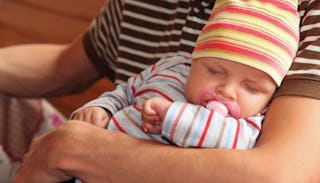 A new dad wearing a brown and white striped t-shirt holding his baby who’s wearing a striped hat and...