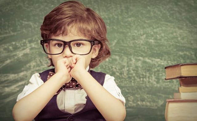 A small boy with glasses sitting with a stack of books in front of him and a green wall behind him