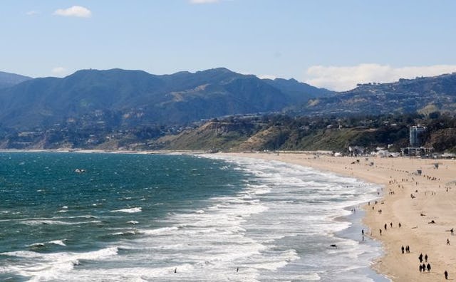 A panorama of a beach in Santa Monica, showing people on the sand beach, a restless sea, and mountai...