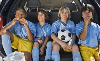 Four boys seated in a van, wearing blue soccer jerseys paired with yellow shorts and blue socks, and...