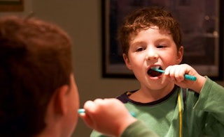A boy in a green shirt brushing his teeth while looking at his reflection in a mirror