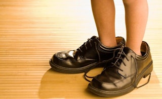 A close-up of a teen's feet wearing an oversized adult man's brown leather shoes