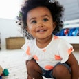 Curly and black-haired baby in a white shirt with peach dots and grey shorts playing on the living r...