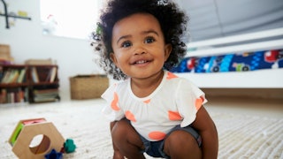 Curly and black-haired baby in a white shirt with peach dots and grey shorts playing on the living r...