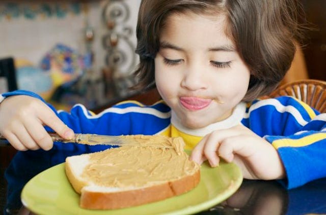 A brown-haired young kid in a white, yellow, and blue shirt smearing peanut butter on a slice of bre...