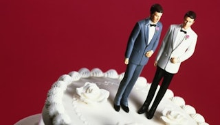 Two groom figurines on top of a white wedding cake with a burgundy background 