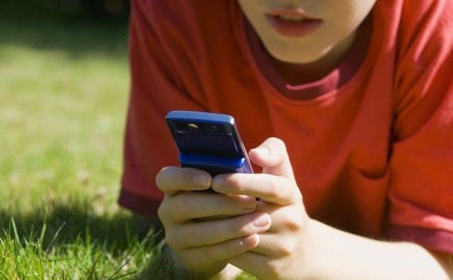 A teen lying on the grass in a red t-shirt while texting on his phone while wearing an orange T-shir...