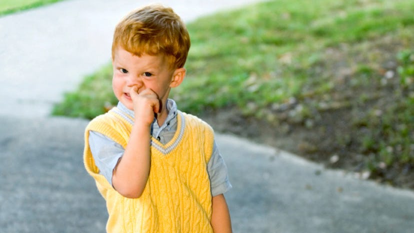 A little kid in a yellow sweater and grey shirt picking his nose.