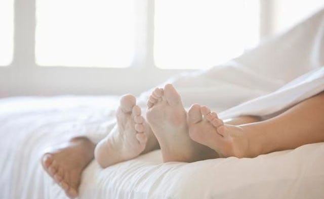 Two persons lying on a bed, covered with a white sheet, with their feet extending beyond the covers.