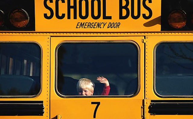 A blonde boy waving through the back window of a yellow bus driving in the distance on his way to th...