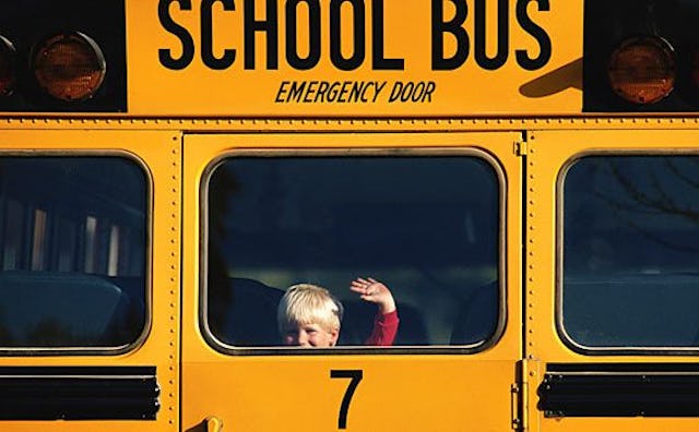 A blonde boy waving through the back window of a yellow bus driving in the distance on his way to th...