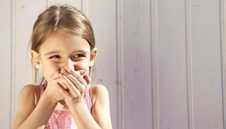 A kid holding her hands over her mouth and smiling in front of a white wall