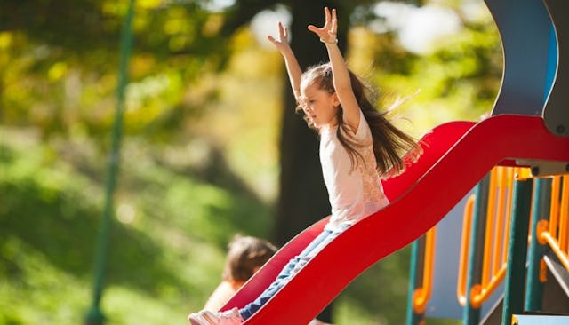 A child with Down Syndrome sliding down a red slide in a park with both arms raised in the air