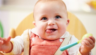 A baby smiling and playing with a spoon while having a meal
