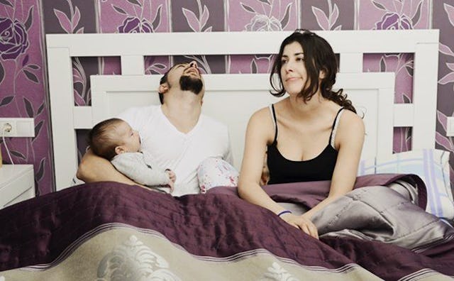 A husband sleeping in a bed while holding his baby and his wife sitting next to them