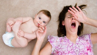 A yawning dark-haired woman lying on the floor in a pink patterned top with a baby in diapers smilin...