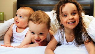 A brunette wavy-haired girl lying in bed next to two toddlers in white tank tops in white bed sheets