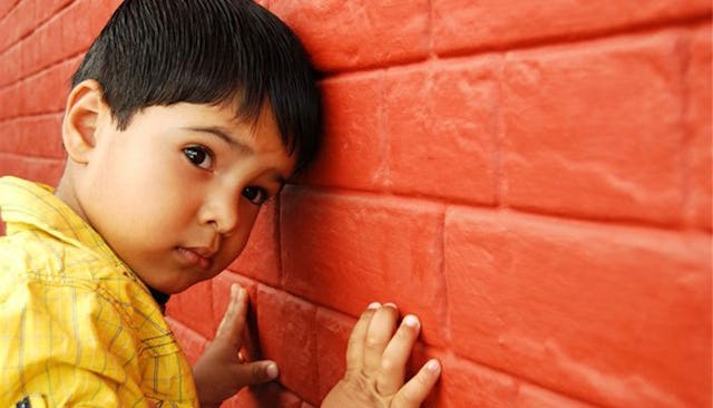 A boy with autism in a yellow shirt leaning his head and hands on a red wall