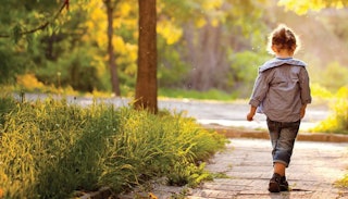 A little girl in a grey jacket and blue denim jeans walking down a side path next to a road surround...