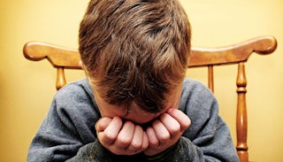 A boy in with his hands on his face crying over the death of a friendship on a wooden chair 