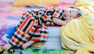 A happy little girl wearing checked pajamas, lying down on her bed with yellow-blue-pink sheets