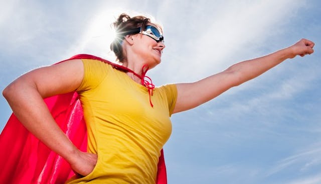 A woman in a yellow shirt, sunglasses and a red cape posing like Superman with one arm stretched out