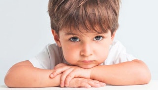 A male kid with brown hair sitting and looking into the camera with a sad facial expression