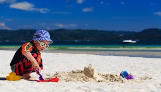 A baby boy wearing a blue checkered hat, white sunglasses, and a colorful shirt makes sand castles a...