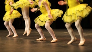 Four little girls wearing yellow tutus and ballet shoes dancing at a dance recital