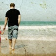 A young father wearing a black t-shirt is holding his son's hand while walking toward the sea during...