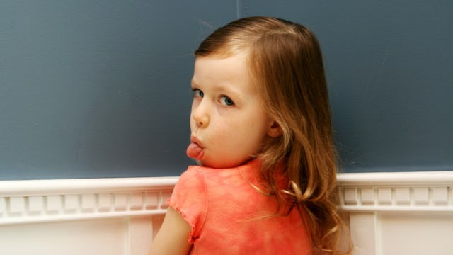 A young child sticking her tongue out while standing in the corner of a room