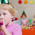 A kid blowing on a pink party whistle with other kids in the background attending the party