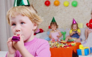 A kid blowing on a pink party whistle with other kids in the background attending the party