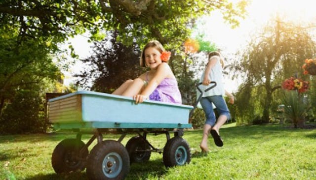 A girl pulling a wheelbarrow with another girl in it in a garden