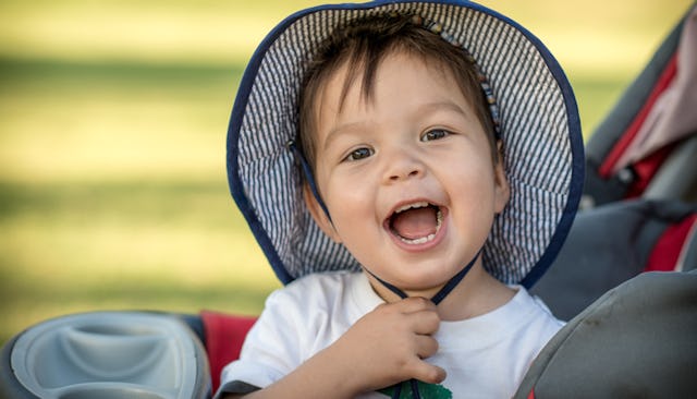 A brown-haired, brown-eyed toddler boy with a hat on his head, sitting in a stroller, smiling