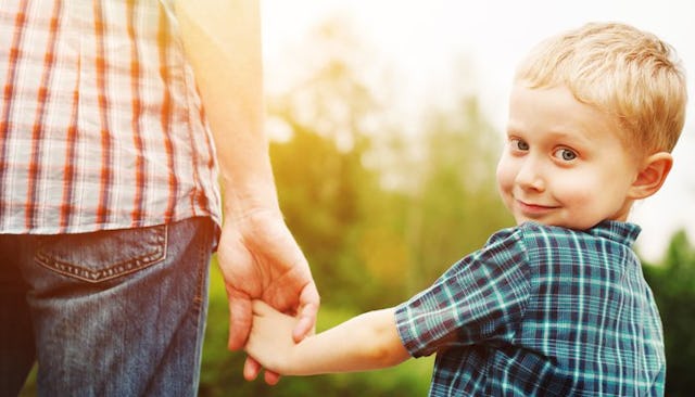 A small young boy with blue eyes and a blue shirt holding his mom's hand while standing