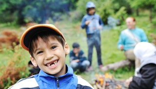 A close-up of a boy smiling at an overnight camp with his family blurred in the background next to a...