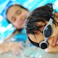 Little kid wearing swim goggles in a pool, smiling, girl in a blurry background