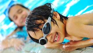 Little kid wearing swim goggles in a pool, smiling, girl in a blurry background