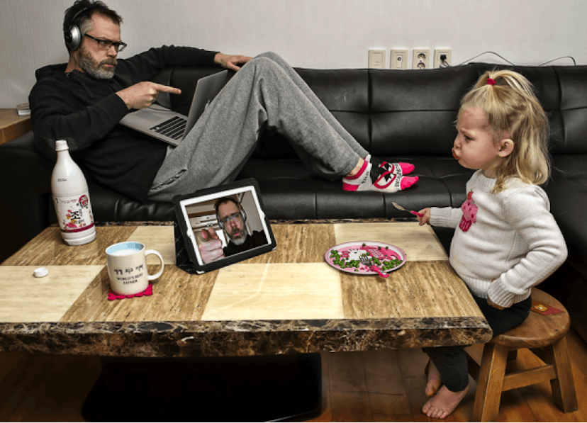 Father with headphones on looking and pointing at his daughter through the web camera while she has ...