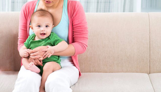 A blue-eyed baby boy in a green bodysuit smiling, sitting on his mother's lap on the couch