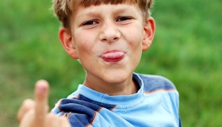 A little boy in a blue shirt showing his middle finger and sticking his tongue out 