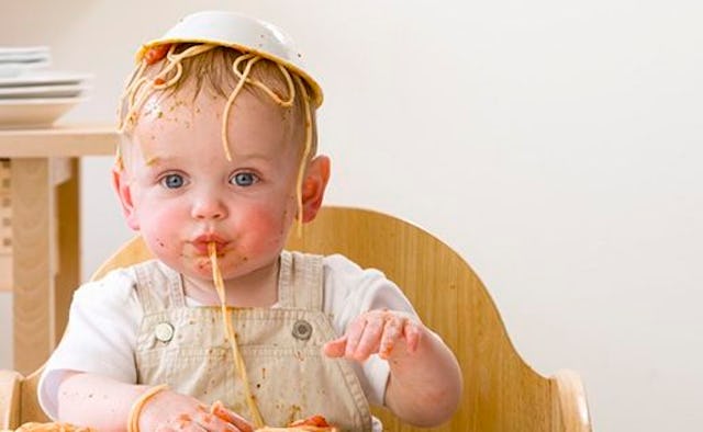 A baby in a restaurant in a white T-shirt and beige rompers with a bowl on its head and spaghetti an...