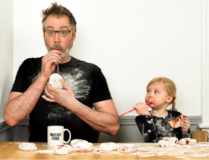 Father and daughter eating and playing with straws and jam-filled donuts on a wodden table with the ...