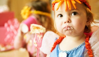 A ginger toddler girl dressed up as a fairy with an orange flower band on her head, messily eating a...
