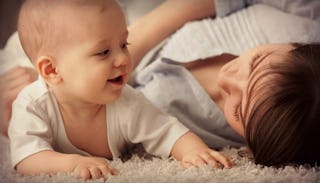 A smiling baby in a white T-shirt crawling on the floor and a mother in a white shirt lying next to ...