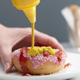  A woman's hand putting mustard on a donut that's already covered with ketchup